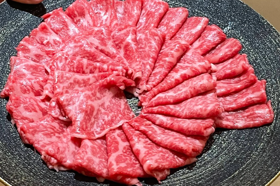 A top-down view of a round, dark textured platter filled with numerous thin slices of raw, high-grade Wagyu beef. The beef is arranged in a decorative, overlapping circular fan pattern. The meat is a vibrant pinkish-red with dense, intricate white marbling (intramuscular fat) throughout. The arrangement fills nearly the entire frame, highlighting the quality and texture of the beef intended for shabu-shabu or yakiniku.