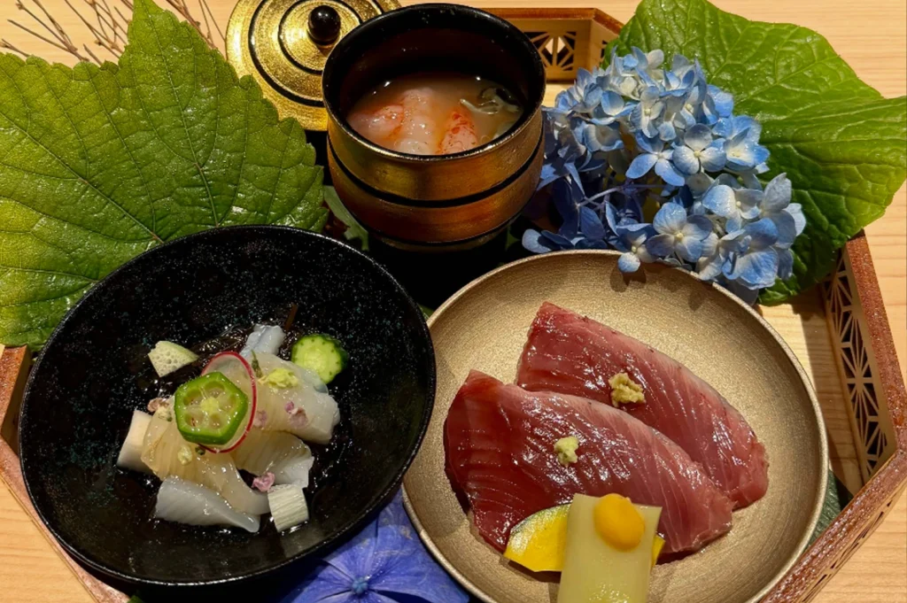 A high-angle, top-down food photograph showcases an exquisite, seasonally themed Japanese omakase course arranged beautifully on a light wooden tray. The artistic presentation features three distinct dishes nestled among large, textured green leaves and a vibrant cluster of blue and purple hydrangea blossoms. In the lower-left, a matte black bowl contains delicate, translucent slices of white fish or squid, intricately garnished with a star-shaped okra slice, a thin radish ring, and tiny pink blossoms. To the right, a shallow bronze-metallic plate displays two thick, glossy slices of deep-red sashimi topped with a dab of grated ginger and paired with small, colorful vegetable accents. At the top center, a black and gold lacquered cup holds a clear soup featuring a plump pink shrimp, with its ornate golden lid resting nearby, completing a luxurious and meticulously crafted dining experience.