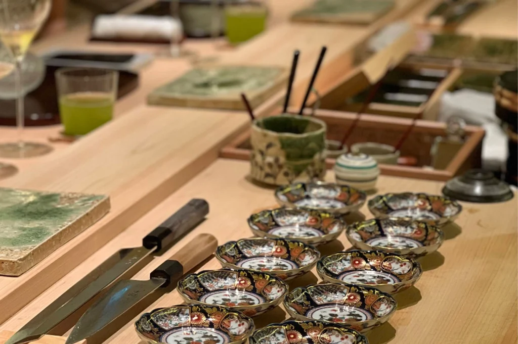 A detailed shot of the Kappo counter prep area featuring two professional Japanese chef knives (yanagiba) resting on the wood. A row of ornate, gold-rimmed ceramic dipping bowls with floral patterns is lined up next to them. In the blurred background, glasses of green tea and wine indicate an active dining service.