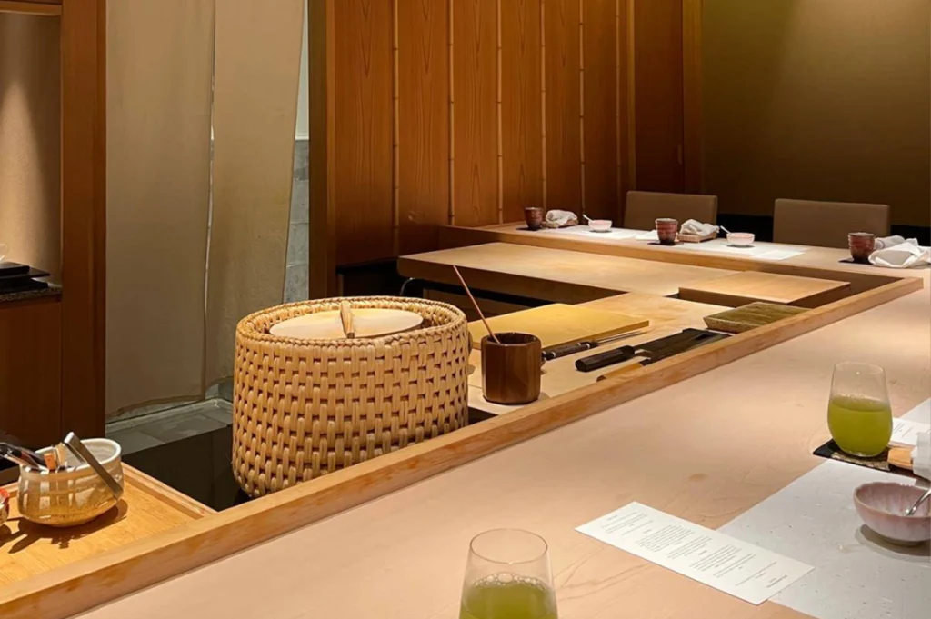 A high-angle, medium shot of a sushi preparation area from the diner's perspective. The light wood counter is meticulously organized, featuring a large woven lidded basket for rice (shari), several professional sushi knives (yanagiba) resting on a wooden block, and small ceramic bowls containing condiments. Two glasses of vibrant green tea sit on the counter. In the background, additional place settings are visible, separated by a vertical wooden slat partition, maintaining a sense of privacy and focused culinary theater.
