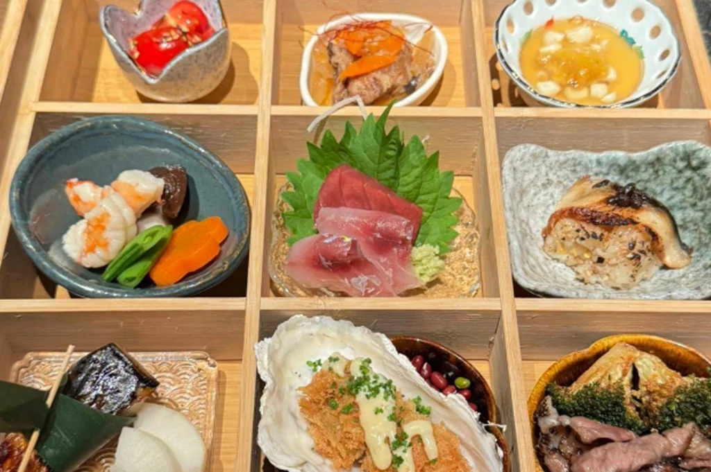 A top-down, close-up view of a wooden bento-style box divided into nine square compartments, each containing a different small dish. The variety includes items such as sashimi on a shiso leaf, braised fish, cooked vegetables, a grilled rice dish, and a breaded oyster, showcasing a curated selection of Japanese culinary preparations.