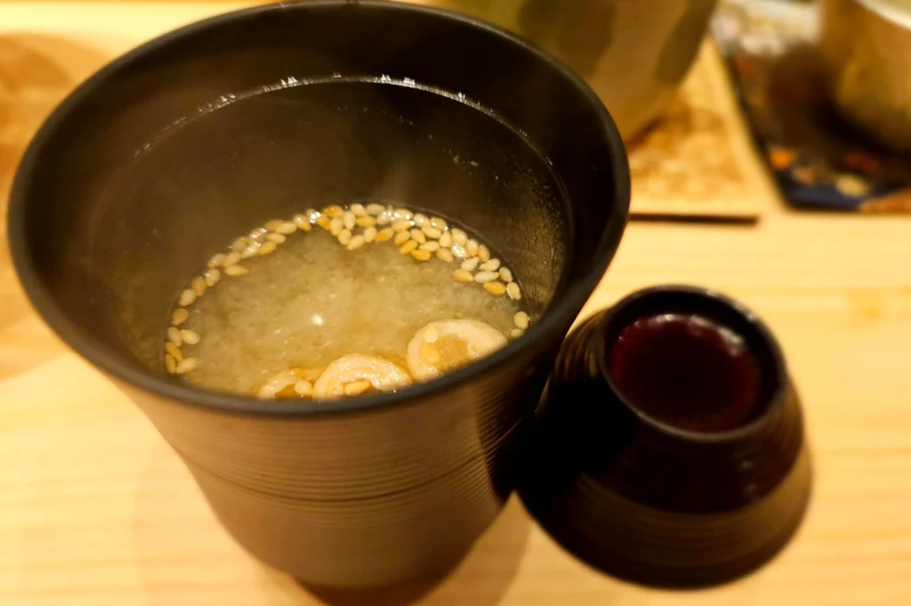 A top-down, slightly blurred close-up of a tall, dark brown textured ceramic cup containing warm miso soup. Floating on the surface are several toasted sesame seeds and a few small, circular pieces of fu (wheat gluten). To the right sits the matching ceramic lid. The background shows the soft, warm tones of the wooden sushi counter, evoking the comforting conclusion of a meal.