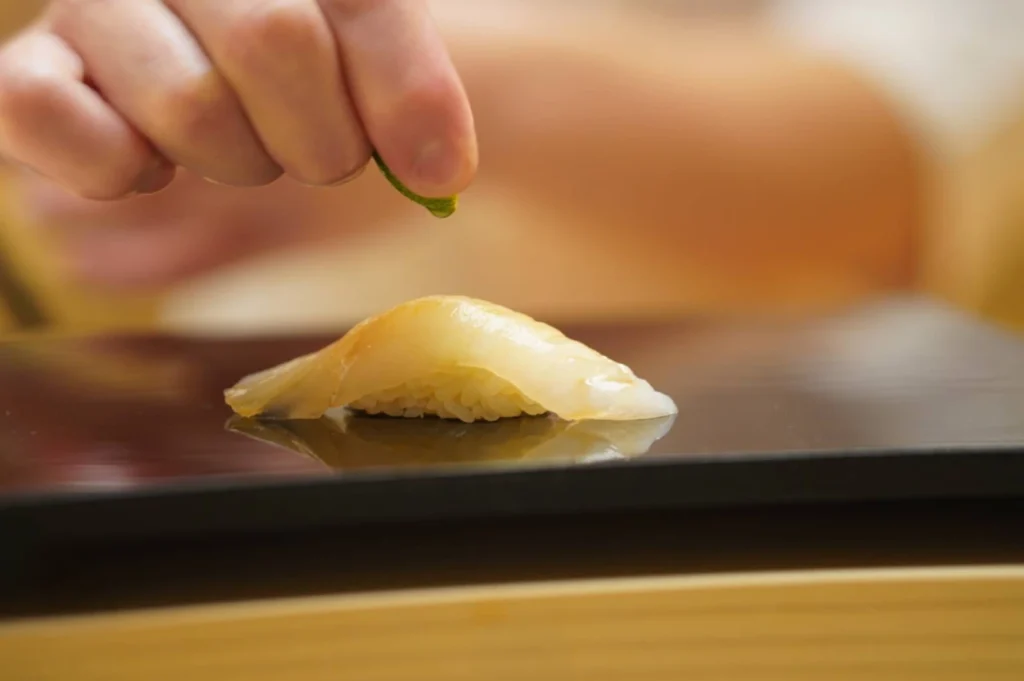 A close-up shot captures a chef’s hand squeezing a small slice of lime over a single piece of nigiri sushi placed on a sleek, reflective black plate. The nigiri features a translucent, pale white fish slice neatly draped over a small bed of seasoned rice, with its form perfectly mirrored on the glossy surface below. The chef's hand is in sharp focus as it holds the lime wedge just above the fish, while the background is softly blurred into warm, out-of-focus tones of beige and cream, emphasizing the precise, artistic preparation of the dish.