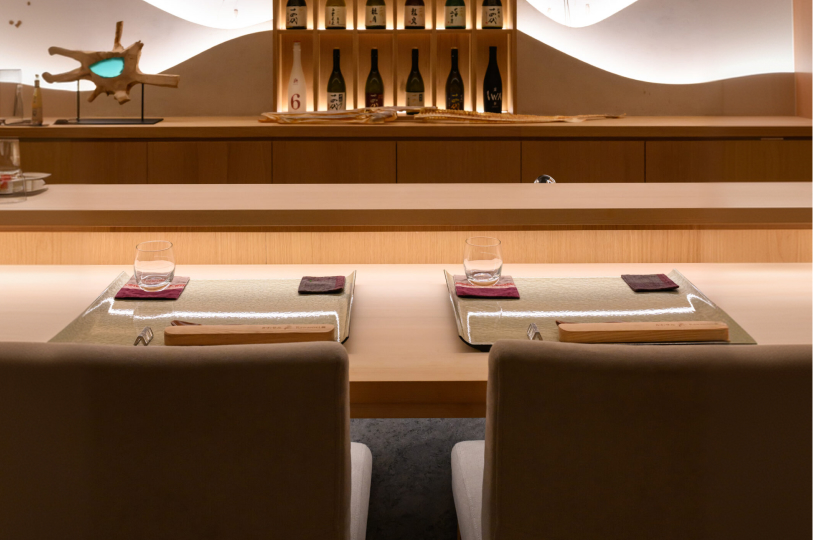 A minimalist sushi counter featuring two elegant place settings with glasses and wooden utensil holders, set against a background of sake bottles and a glowing wave-patterned wall.