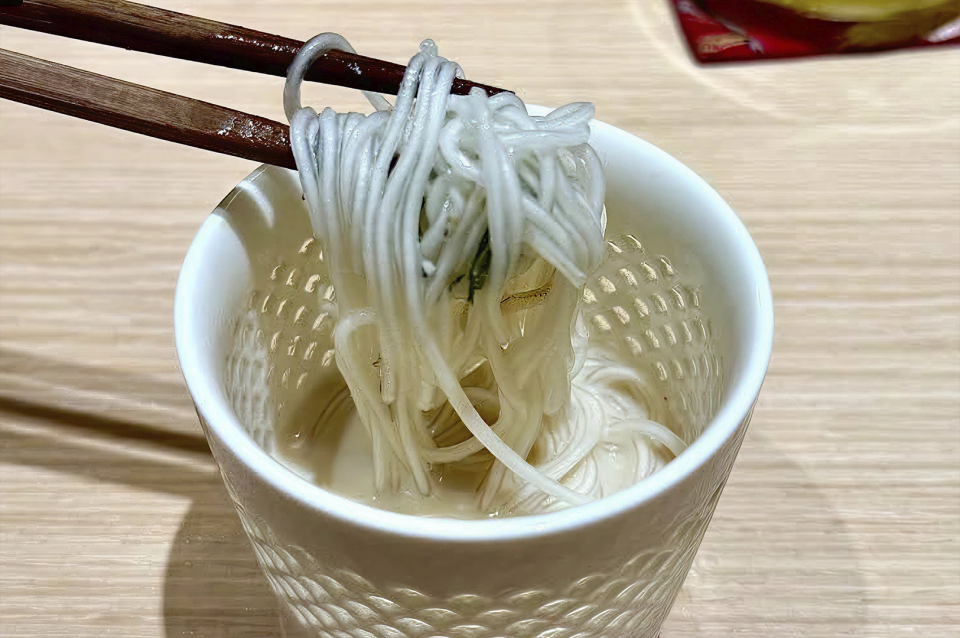 A close-up shot of thin, white noodles somen being lifted from a white textured ceramic cup using dark wooden chopsticks. The noodles are coated in a creamy, translucent white broth or sauce. The cup features a decorative scalloped or "raindrop" texture on its exterior. The scene is set against a light wood tabletop, capturing a moment of a meal being enjoyed.