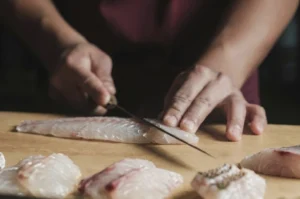 A close-up shot of a chef’s hands skillfully slicing a fresh sea bass on a wooden cutting board. The chef uses a long, slender Japanese-style knife to make a precise cut through the translucent, pearly-white flesh. Several neatly portioned pieces of sea bass, some showing the delicate skin, are arranged in the soft-focus foreground.