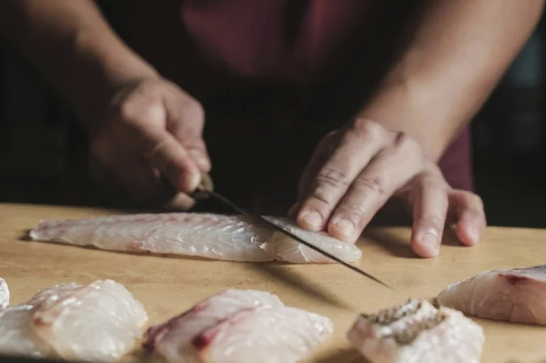 A close-up shot of a chef’s hands skillfully slicing a fresh sea bass on a wooden cutting board. The chef uses a long, slender Japanese-style knife to make a precise cut through the translucent, pearly-white flesh. Several neatly portioned pieces of sea bass, some showing the delicate skin, are arranged in the soft-focus foreground.