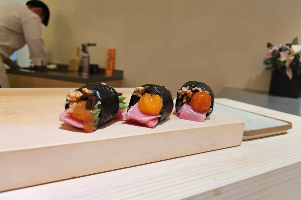 A close-up, eye-level shot captures three meticulously crafted sushi handrolls, known as maki, lined up horizontally on a light-colored wooden serving block. Each roll is wrapped in dark, glossy nori seaweed and overflows with a vibrant assortment of ingredients: thick slices of pale pink fatty tuna (otoro), a bright orange cured egg yolk, dark simmered mushrooms, and a sprinkle of toasted rice pearls for texture. The sushi sits on a clean, minimalist wooden counter that leads the eye toward a soft-focus background where a sushi chef in a white uniform works in a modern, brightly lit kitchen. To the right, a small dark vase with white and pink flowers adds a touch of elegance to the serene, high-end dining atmosphere.