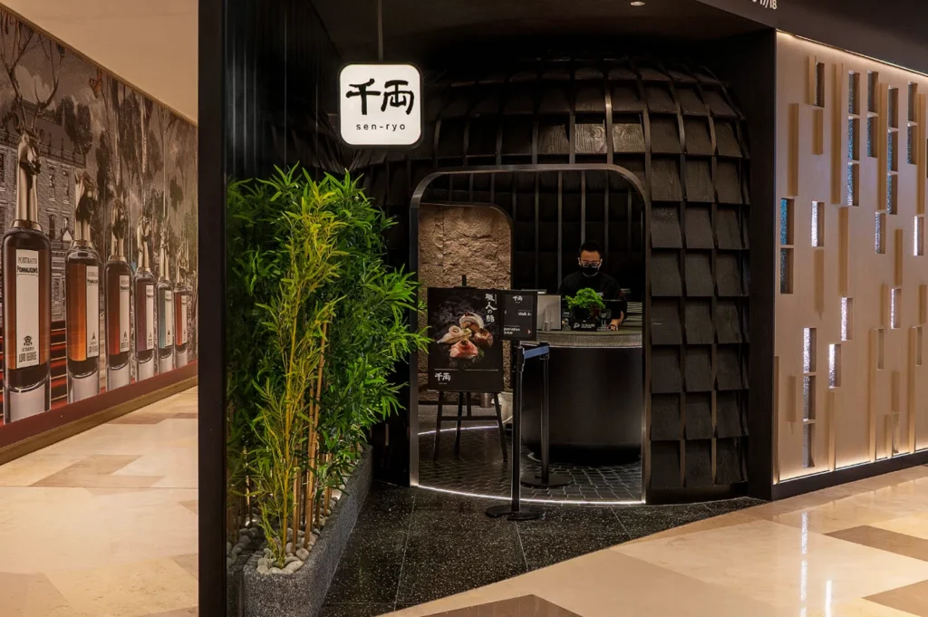 The sleek, modern entrance of a Sen-ryo restaurant located within a brightly lit mall corridor. The storefront features a unique, dark architectural structure resembling a shingle-textured dome or hut, with a circular entryway where a masked host stands behind a black reception podium. To the left, a tall cluster of faux bamboo sits in a stone planter against a wall adorned with large-scale artwork of perfume bottles, while the restaurant’s glowing white sign with Japanese calligraphy hangs prominently above the entrance.