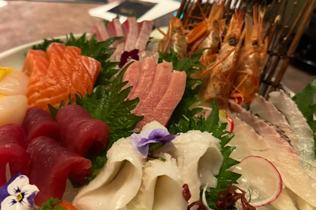 A vibrant, close-up shot of a lavish sashimi platter featuring a variety of fresh seafood. The arrangement includes bright orange salmon, deep red tuna, delicate white fish, and plump scallops, all nestled against green shiso leaves and garnished with small purple and white edible flowers. In the upper right, several whole spot prawns with long antennae are artfully displayed against a rustic wooden backdrop, while thin slices of radish add a crisp detail to the foreground.