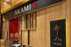 A wide shot of the Akami restaurant entrance. The facade features light-toned vertical wood paneling and a large black header with "AKAMI" in white capital letters. A traditional red noren (divided curtain) with a white wave pattern hangs over the entrance, and a large black textured sign on the right displays the restaurant’s name in both golden Japanese calligraphy and white English text. A small wooden sign hanging by the door indicates that the restaurant is open for business.