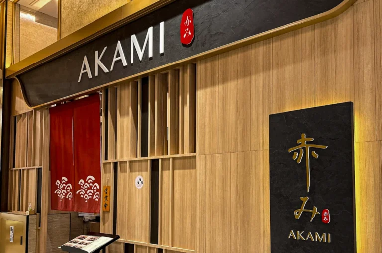 A wide shot of the Akami restaurant entrance. The facade features light-toned vertical wood paneling and a large black header with "AKAMI" in white capital letters. A traditional red noren (divided curtain) with a white wave pattern hangs over the entrance, and a large black textured sign on the right displays the restaurant’s name in both golden Japanese calligraphy and white English text. A small wooden sign hanging by the door indicates that the restaurant is open for business.