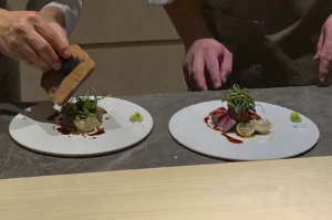 A close-up, high-angle shot of two chefs in white uniforms meticulously plating a gourmet dish on a grey stone counter. Two identical white circular plates feature a central arrangement of seared beef or tuna medallions topped with a nest of thin green scallion slivers and white onion bulbs, drizzled with a dark balsamic or soy-based glaze. A small dollop of bright green wasabi sits on the edge of each plate. One chef’s hand is visible using a wooden tool to press or finish a component of the dish.