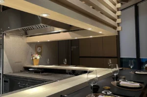 A wide-angle shot of a modern, upscale Omakase restaurant interior in Singapore. A sleek, L-shaped light wood counter dominates the foreground, set with black placemats, wine glasses, and ceramic tea cups. Above the counter, a large, geometric architectural feature made of stacked light wood beams houses recessed spotlights that cast a warm glow. In the background, a professional stainless steel kitchen with a large vent hood is visible behind a stone-textured backsplash.