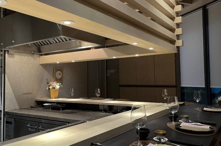 A wide-angle shot of a modern, upscale Omakase restaurant interior in Singapore. A sleek, L-shaped light wood counter dominates the foreground, set with black placemats, wine glasses, and ceramic tea cups. Above the counter, a large, geometric architectural feature made of stacked light wood beams houses recessed spotlights that cast a warm glow. In the background, a professional stainless steel kitchen with a large vent hood is visible behind a stone-textured backsplash.
