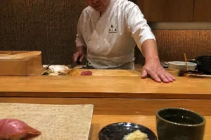 An eye-level shot captures a sushi chef in a clean white uwagi (traditional jacket) standing behind a tiered light-wood counter. His left hand is planted firmly on the lower service ledge, while his right hand holds a professional knife poised over a single, vibrant piece of raw tuna on a wooden cutting board. The background features a textured, earthy-toned wall and minimalist kitchen shelves. In the foreground, from the diner's perspective, a piece of finished nigiri sits on a stone platter to the left, accompanied by a small dish of pickled ginger and a dark ceramic cup of tea on the right, creating a sense of intimate, focused craftsmanship.