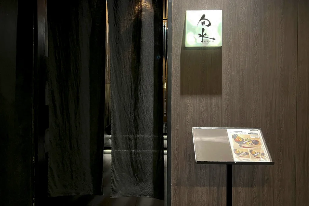 The entrance to Shunsui, a Japanese restaurant. The doorway is obscured by heavy, dark charcoal-grey noren (traditional fabric dividers), which are parted slightly to reveal a glimpse of a warmly lit interior. To the right of the drapes, a square, backlit glass sign features minimalist black Japanese calligraphy. Below the sign, a sleek black menu stand displays a printed menu with vibrant food photography. The surrounding wall is composed of dark, vertical wood grain panels, creating a sophisticated and private atmosphere.