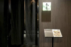 The entrance to Shunsui, a Japanese restaurant. The doorway is obscured by heavy, dark charcoal-grey noren (traditional fabric dividers), which are parted slightly to reveal a glimpse of a warmly lit interior. To the right of the drapes, a square, backlit glass sign features minimalist black Japanese calligraphy. Below the sign, a sleek black menu stand displays a printed menu with vibrant food photography. The surrounding wall is composed of dark, vertical wood grain panels, creating a sophisticated and private atmosphere.
