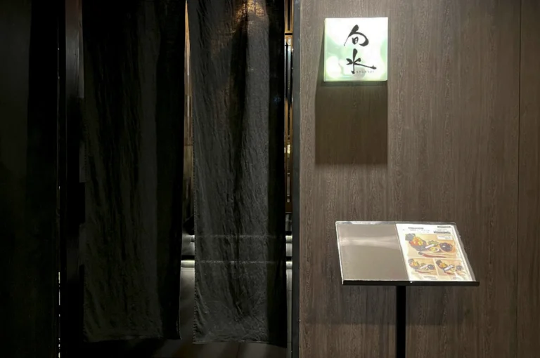 The entrance to Shunsui, a Japanese restaurant. The doorway is obscured by heavy, dark charcoal-grey noren (traditional fabric dividers), which are parted slightly to reveal a glimpse of a warmly lit interior. To the right of the drapes, a square, backlit glass sign features minimalist black Japanese calligraphy. Below the sign, a sleek black menu stand displays a printed menu with vibrant food photography. The surrounding wall is composed of dark, vertical wood grain panels, creating a sophisticated and private atmosphere.