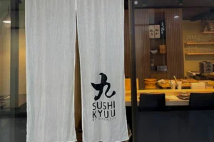 A storefront view of Sushi Kyuu by Shunsui located at Orchard Plaza. A long, semi-transparent white linen noren (traditional Japanese fabric divider) hangs at the entrance, featuring a large black calligraphic character above the restaurant's name. Through the glass door and the gap in the curtains, the warmly lit interior is visible, showcasing a light wood sushi counter, black minimalist chairs, and a back wall with wooden shelving holding sake bottles and glassware.