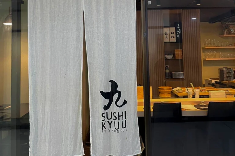 A storefront view of Sushi Kyuu by Shunsui located at Orchard Plaza. A long, semi-transparent white linen noren (traditional Japanese fabric divider) hangs at the entrance, featuring a large black calligraphic character above the restaurant's name. Through the glass door and the gap in the curtains, the warmly lit interior is visible, showcasing a light wood sushi counter, black minimalist chairs, and a back wall with wooden shelving holding sake bottles and glassware.