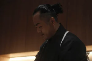 A cinematic, side-profile close-up of a sushi chef with dark hair tied in a neat bun. He is looking down with deep focus, his face partially illuminated by a warm, soft light against a dark, moody wooden background. He wears a traditional dark Japanese chef's uniform with white straps.