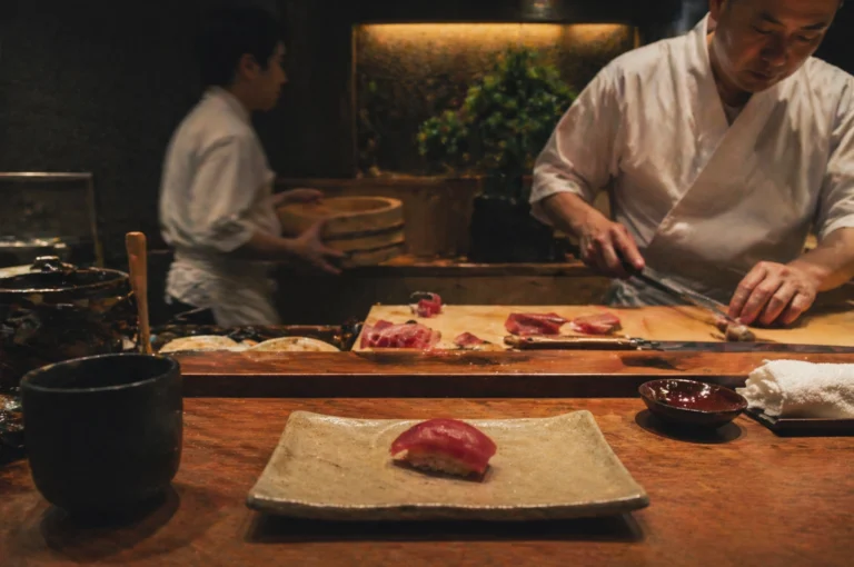 A serene moment at a high-end sushi bar, where a single piece of dark pink tuna nigiri sits centered on a rectangular, textured ceramic plate in the foreground. To the left of the plate is a dark, matte ceramic tea cup, and to the right, a small dish of soy sauce and a neatly folded white hand towel. In the blurred midground, an expert sushi chef in a crisp white chef's coat is meticulously slicing pieces of raw fish on a light wooden cutting board with a long, professional knife. Behind him, another staff member in a white uniform carries a traditional wooden sushi rice tub (hangiri) across the dim, warm-lit kitchen. The background features dark wood accents and a small bonsai tree illuminated by soft, golden light, creating an intimate and disciplined atmosphere.