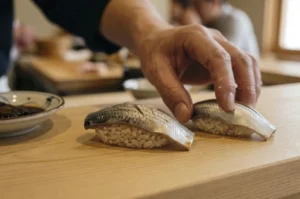 A close-up, shallow-depth-of-field shot captures a sushi chef’s hand placing a second piece of Kohada (gizzard shad) nigiri onto a light-colored wooden counter. The fish features shimmering, silver-and-black speckled skin with precise decorative cross-hatch incisions, resting atop a small, oblong mound of seasoned sushi rice. The chef's hand is frozen in a delicate motion, with a few stray grains of rice clinging to the thumb, emphasizing the artisanal nature of the preparation. In the soft-focus background, the warm, minimalist interior of a high-end sushi bar is visible, including a small dish of soy sauce to the left and the blurred silhouette of another person seated at the counter.