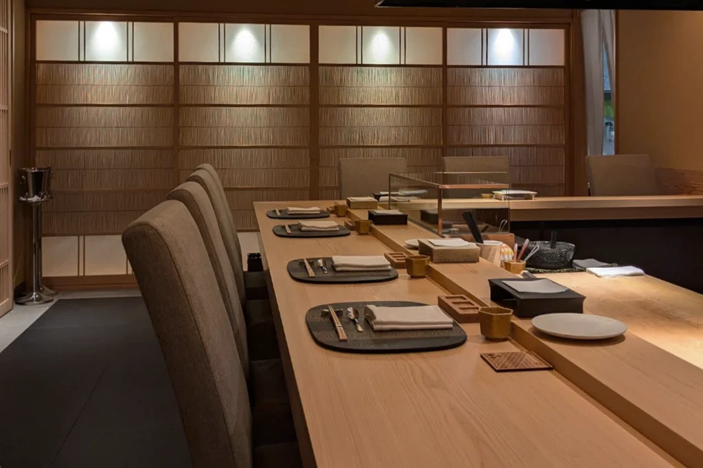 A wide-angle interior shot of a sophisticated, minimalist Japanese Kappo-style dining room. A long, polished light-wood counter dominates the foreground, set with several individual placemats featuring dark textured chargers, white linen napkins, and wooden chopsticks. In the background, floor-to-ceiling shoji-inspired sliding screens with delicate vertical wooden slats create a warm, private atmosphere under soft recessed lighting.