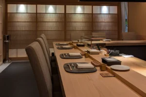 A wide-angle interior shot of a sophisticated, minimalist Japanese Kappo-style dining room. A long, polished light-wood counter dominates the foreground, set with several individual placemats featuring dark textured chargers, white linen napkins, and wooden chopsticks. In the background, floor-to-ceiling shoji-inspired sliding screens with delicate vertical wooden slats create a warm, private atmosphere under soft recessed lighting.