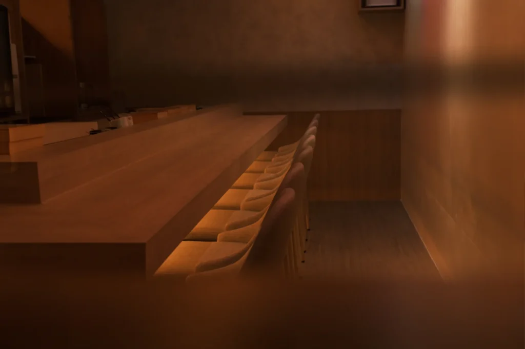 A perspective shot of a serene, empty sushi bar, showing a polished wooden counter with several chairs lined up, illuminated by soft, warm, indirect lighting that creates a cozy and intimate atmosphere.