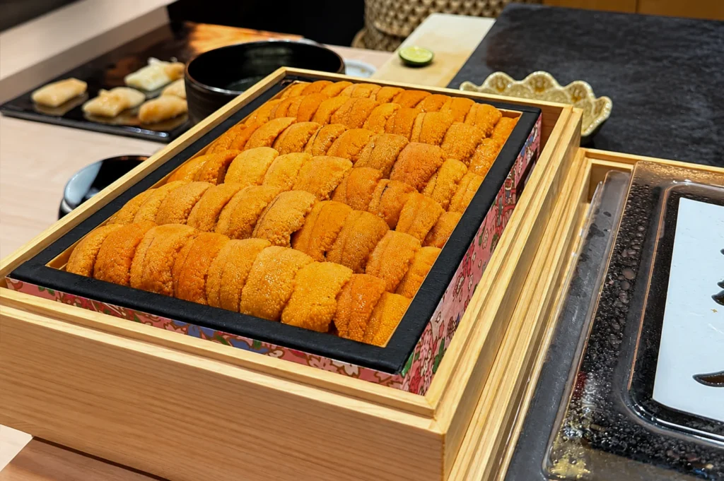 A wooden box filled with a generous portion of fresh, bright orange sea urchin (uni) arranged in neat, side-by-side rows, resting on the sushi bar counter with a hint of other prepared sushi visible in the blurred background.