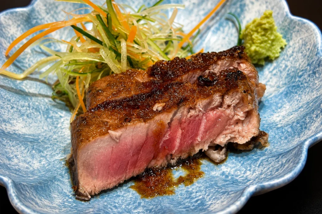 A seared piece of steak, cooked to a medium-rare pink center with a dark, flavorful crust, is served on a light blue, flower-shaped bowl alongside a small mound of shredded cucumber and carrot salad and a dollop of bright green wasabi.