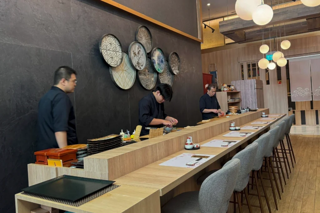 The interior of the restaurant featuring a long, light-wood sushi counter with grey upholstered high-back chairs. Behind the counter, three chefs in black uniforms are meticulously preparing food. The back wall is a dark, slate-grey stone decorated with several circular, patterned woven plates in various sizes and colors. Modern, spherical white pendant lights hang from a rustic wooden ceiling beam, casting a warm glow over the dining area.
