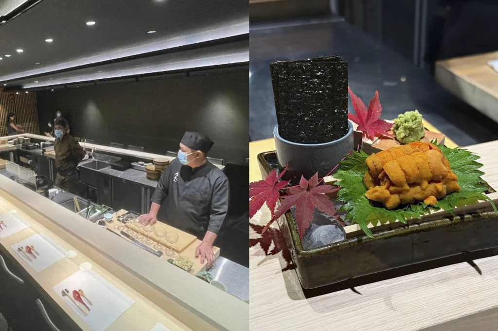 A split-screen image. The left side shows a wide-angle view of a modern, minimalist sushi bar with dark walls and light wood counters; two chefs wearing black uniforms and blue surgical masks stand behind the prep station. The right side is a vibrant close-up of a premium appetizer: a generous portion of bright orange sea urchin (uni) served on a green shiso leaf, nestled in a dark ceramic tray with ice, garnished with red maple leaves and a side of fresh wasabi and crisp nori seaweed sheets.