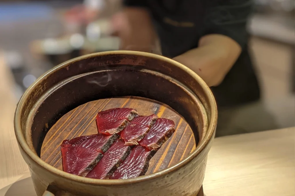 A close-up, top-down view of six thick slices of deep red sashimi, likely bonito or tuna, arranged on a circular wooden platform inside a rustic ceramic pot. Wisps of white smoke curl up from the wood, indicating a cold-smoking process. The fish has a seared, dark outer edge and a glistening, succulent interior. The background is softly blurred, showing the hands of a chef in a dark uniform.