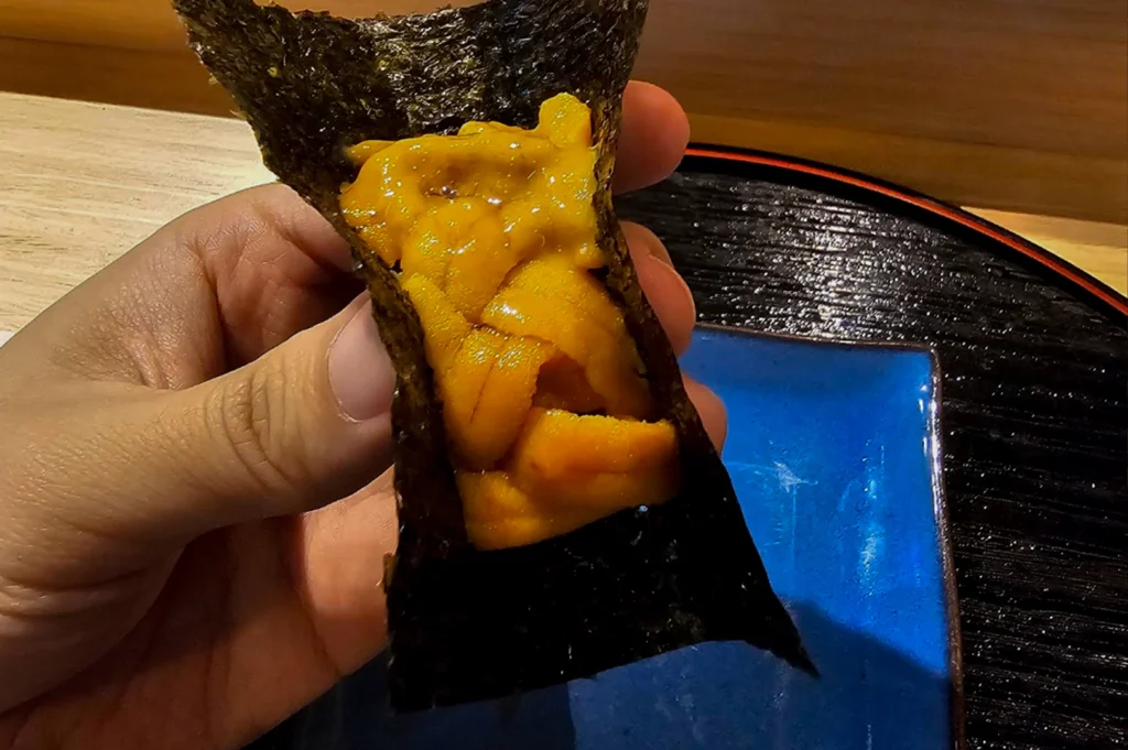 A first-person perspective of a hand holding a premium Uni (sea urchin) handroll. The vibrant, orange lobes of fresh uni are piled generously inside a crisp, dark green sheet of nori (seaweed). The background shows a light wooden table surface and a glimpse of a blue ceramic plate resting on a black lacquered tray with a red rim.