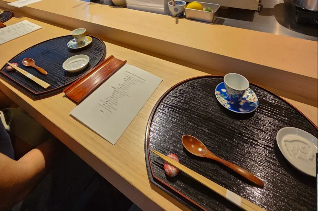 An eye-level, high-angle shot depicts two empty place settings at a light-colored wooden sushi bar, each arranged on an oval, textured black tray. On the right, a small white teacup with blue and floral accents sits on a matching blue saucer, alongside a wooden spoon and a pair of chopsticks resting on a small, smooth, reddish-marbled chopstick rest, with a small white dish nearby. A nearly identical setup is arranged on the left tray, with a printed menu placed between the two settings on the wooden counter surface. In the background, out-of-focus elements include a small blue-and-white cup containing a utensil and a container holding a yellow lemon wedge and a green lime wedge, all resting on a thin, elevated shelf along the back of the sushi bar.