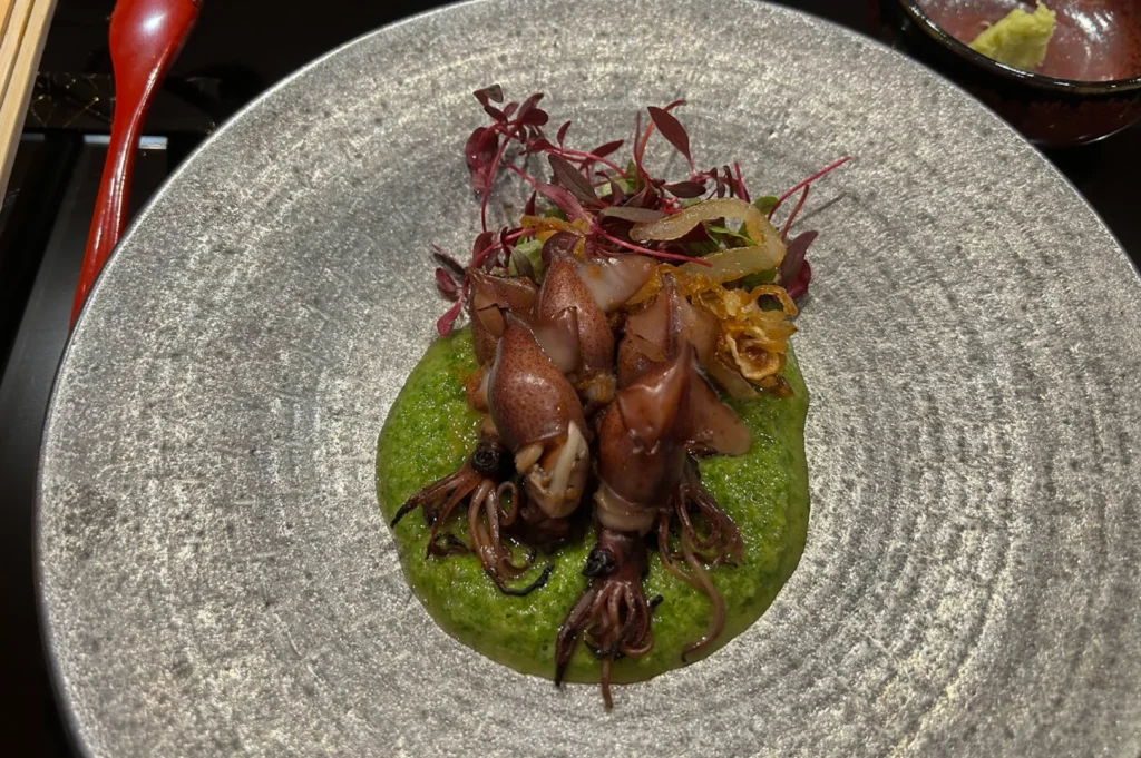 A gourmet appetizer featuring several small, cooked squid arranged atop a vibrant green purée on a textured, grey plate, garnished with a small pile of red microgreens and fried crispy bits.