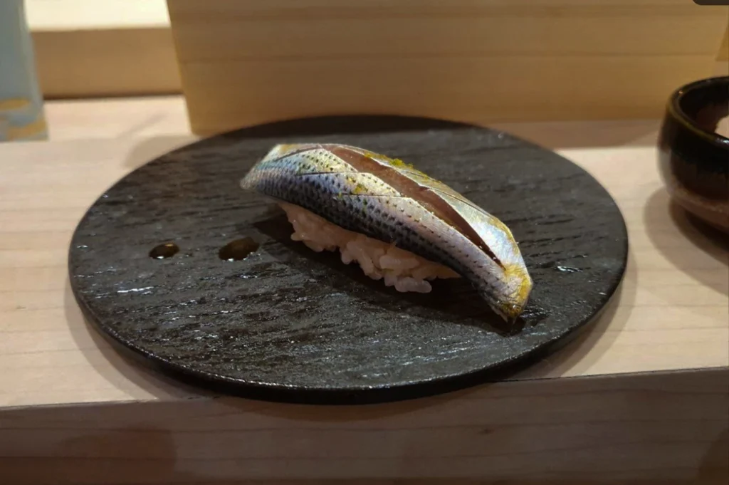 A close-up, eye-level shot highlights a single piece of kohada (gizzard shad) nigiri resting on a round, textured black ceramic plate. The silver-and-blue skinned fish is meticulously scored with decorative crisscross cuts along its spine, subtly glistening with a delicate brush of soy glaze and tiny flecks of green citrus zest. The sushi sits at a slight angle on its compact bed of seasoned rice, positioned on a light wooden sushi counter with a blurred wooden background and two small droplets of sauce accenting the plate.