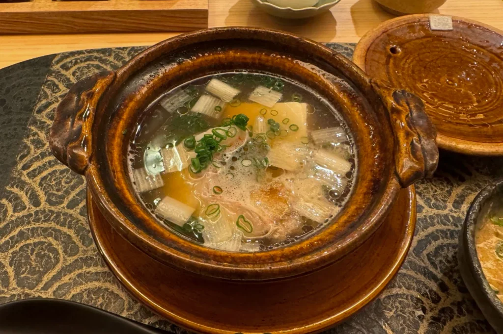 A close-up, top-down view of a Donabe (clay pot) containing a clear, simmering dashi broth. Inside the pot are delicate pieces of white fish, tofu, and vibrant green scallions. The clay pot is a rustic, glazed brown with traditional handles, sitting on a matching saucer over a dark, patterned countertop. Steam visibly rises from the surface, highlighting the "Kappo" focus on cooked dishes.