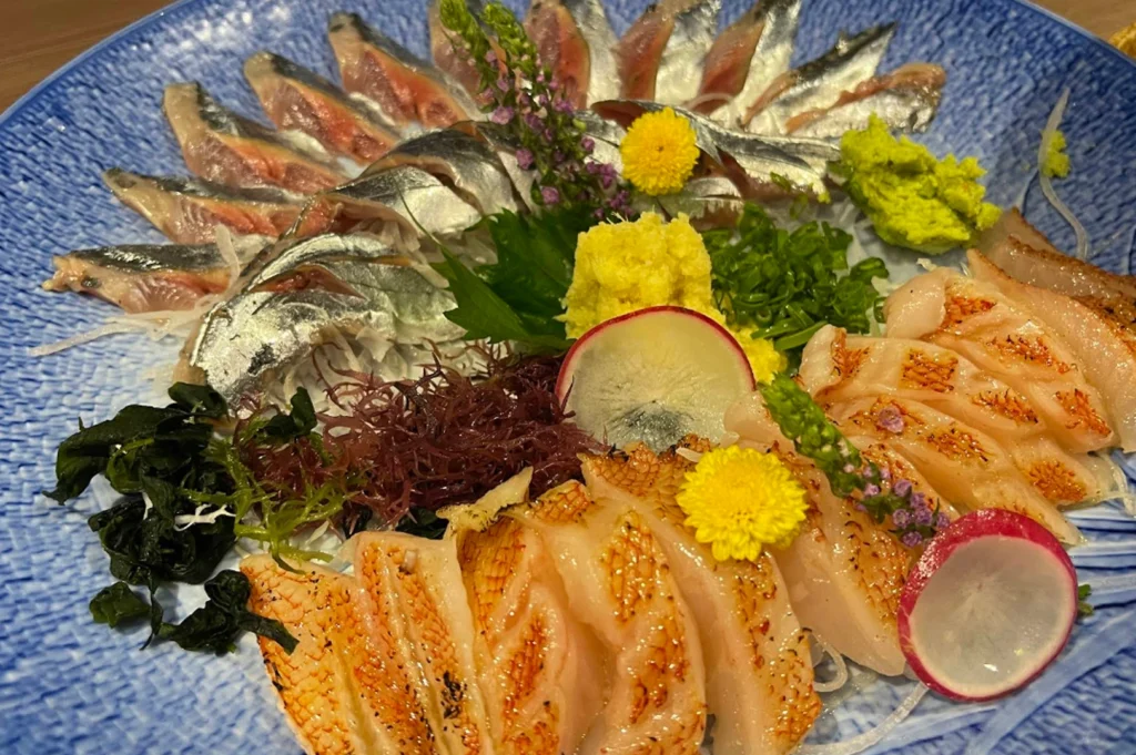 A top-down, close-up shot of a vibrant sashimi platter served on a textured blue ceramic plate. The arrangement includes thinly sliced, silver-skinned fish (likely mackerel or sardine) fanned out at the top, and seared white fish with charred edges at the bottom. The dish is garnished with bright yellow flowers, purple blossoms, shredded daikon radish, fresh wasabi, sliced red radish, and a mound of grated ginger and green onions in the center.