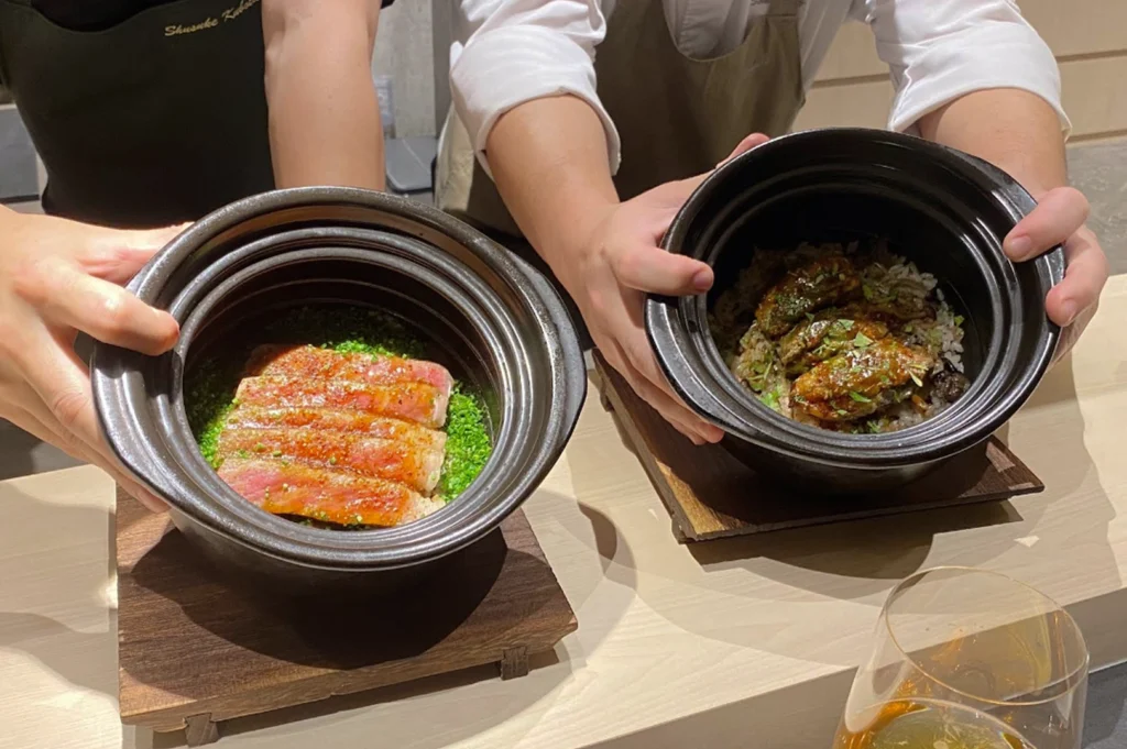 Two chefs in the background hold out two black ceramic donburi bowls toward the camera. The bowl on the left contains sliced, seared wagyu beef topped with orange zest or spice, resting on a vibrant bed of finely chopped green chives. The bowl on the right features grilled eel (unagi) or glazed seafood over seasoned rice with mushrooms and herbs. A glass of amber-colored liquid is partially visible in the bottom right corner.