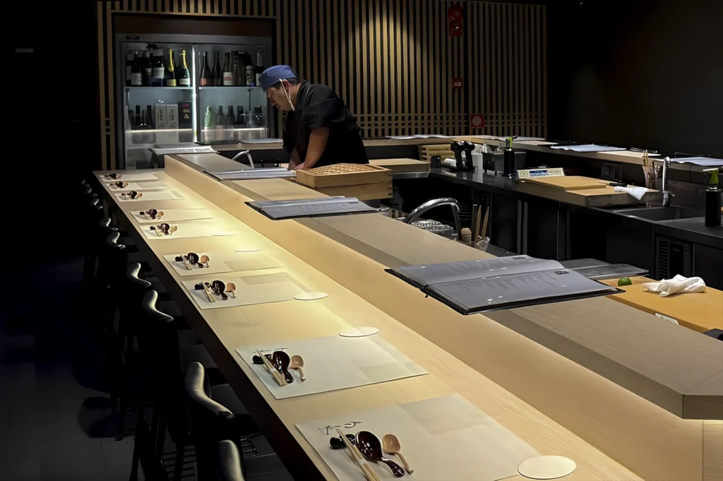 Interior shot of an omakase sushi counter. A chef wearing a black uniform and a blue headwrap is positioned behind the counter, focused on preparation. The long, light-colored wooden counter is impeccably set for multiple guests, with individual place settings featuring dark lacquered spoons and chopsticks on white paper mats. The background features vertical wooden slats and a refrigerated cabinet stocked with sake and champagne. The lighting is focused and warm on the counter surface, while the rest of the room remains in soft shadow.
