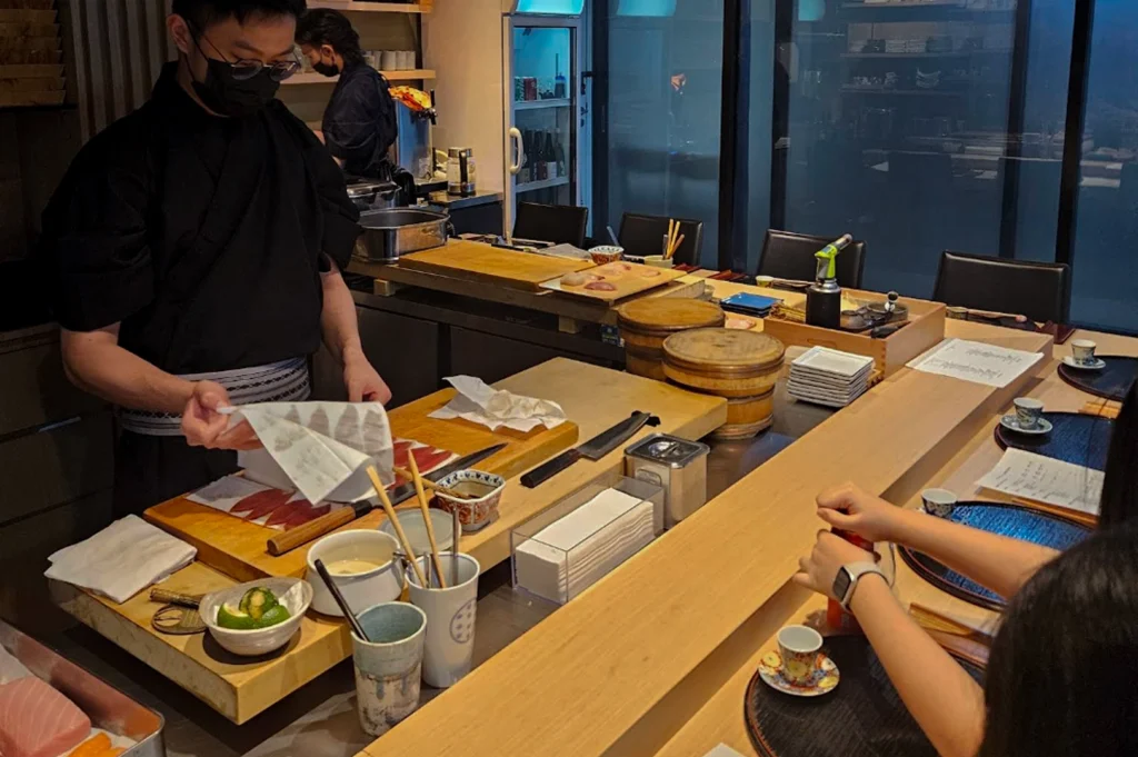An interior shot of the intimate dining area where a sushi chef in a black traditional uniform and face mask is meticulously preparing food behind a light-colored wooden L-shaped counter. The chef is using a white cloth to pat down slices of fresh fish on a wooden cutting board. The counter is set with small tea cups and black circular placemats for guests. In the background, professional kitchen equipment, stacked wooden rice tubs (hangiri), and various condiment containers are neatly organized.