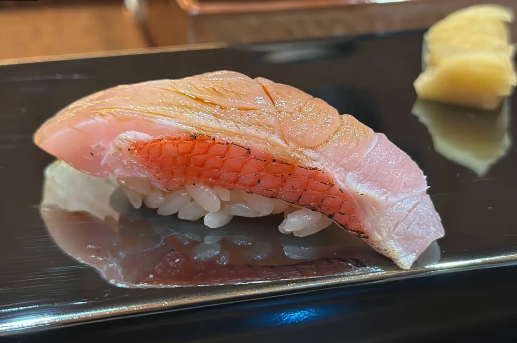 An extreme macro shot of a piece of Kinmedai (Golden Eye Snapper) nigiri. The fish has a beautiful gradient of pale pink to white, with its characteristic bright red skin visible along the side, which appears lightly seared. The flesh is scored with thin diagonal cuts and sits atop a small, tight bed of seasoned white rice. A piece of pickled ginger is blurred in the background on a black lacquer serving board.