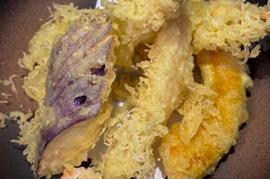 A macro, extreme close-up of golden-brown vegetable tempura. The batter is light, airy, and very crispy, with distinct "lacework" textures. Visible pieces include a slice of purple sweet potato and a piece of orange pumpkin or squash. The lighting emphasizes the delicate, flaky shards of the fried coating.