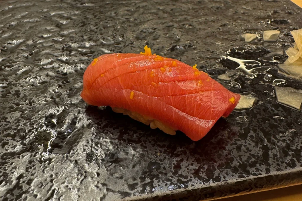 An extreme close-up of a single piece of premium tuna nigiri (chu-toro or akami) served on a textured, dark grey stone plate. The fish is a vibrant pinkish-red with delicate marbling and fine score marks across the top. It is garnished with tiny flakes of bright orange citrus zest. The background of the plate has a wet, obsidian-like sheen with a few small pieces of pickled ginger to the side.
