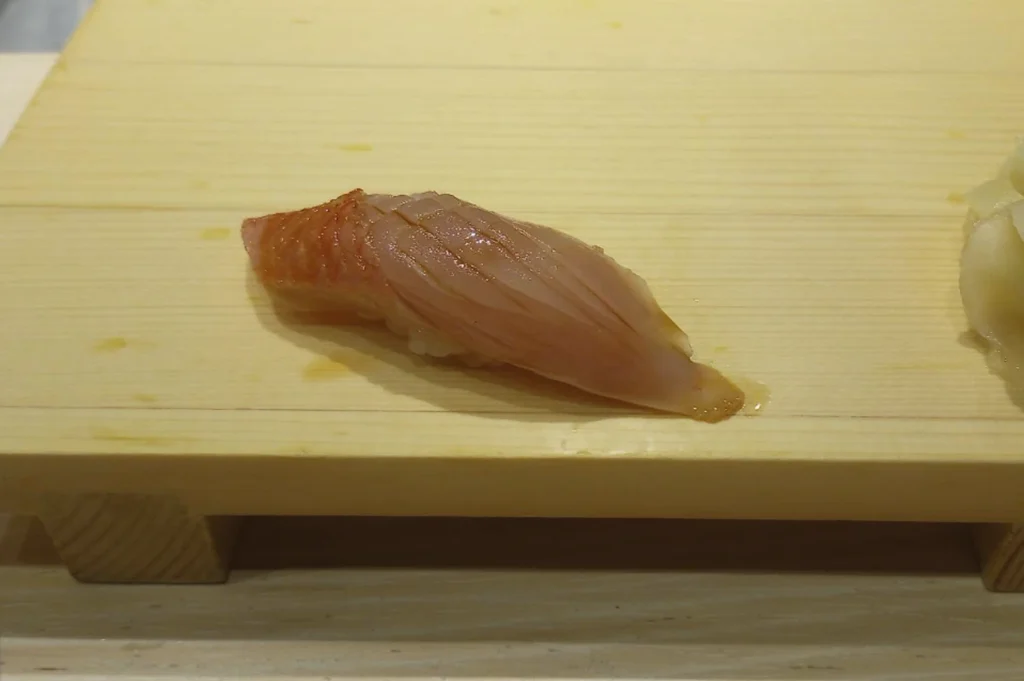 A single piece of Kinmedai (Golden Eye Snapper) nigiri resting on a light-colored wooden sushi board (geta). The fish has a pale pink, translucent flesh with a thin strip of vibrant orange-red skin along the top. Fine, diagonal score marks are visible on the surface of the fish, which has been lightly brushed with a soy-based glaze. A small mound of pickled ginger is visible on the far right edge of the frame. The lighting is soft and even, highlighting the fresh texture of the seafood.