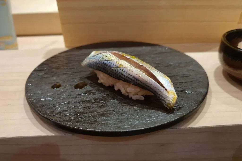 A single piece of nigiri sushi, featuring a slice of silver-skinned fish with decorative cross-hatch scoring on top of a small mound of vinegared sushi rice, presented on a dark, circular stone plate with two small drops of sauce.